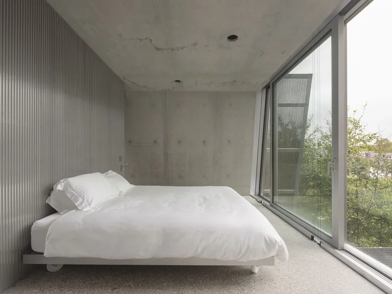 White platform bed under concrete ceiling with floor-to-ceiling glass opening to the garden trees.