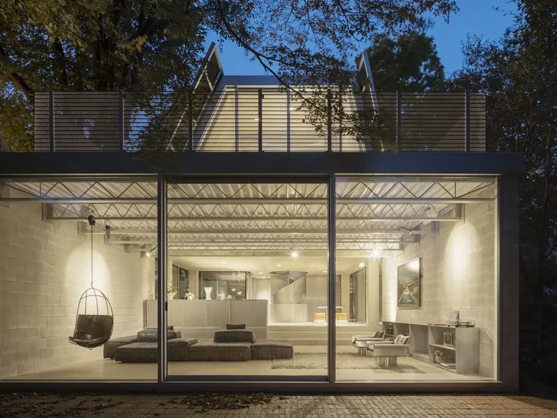The garden facade at night, full-height glass reveals the living area glowing, trusses visible.