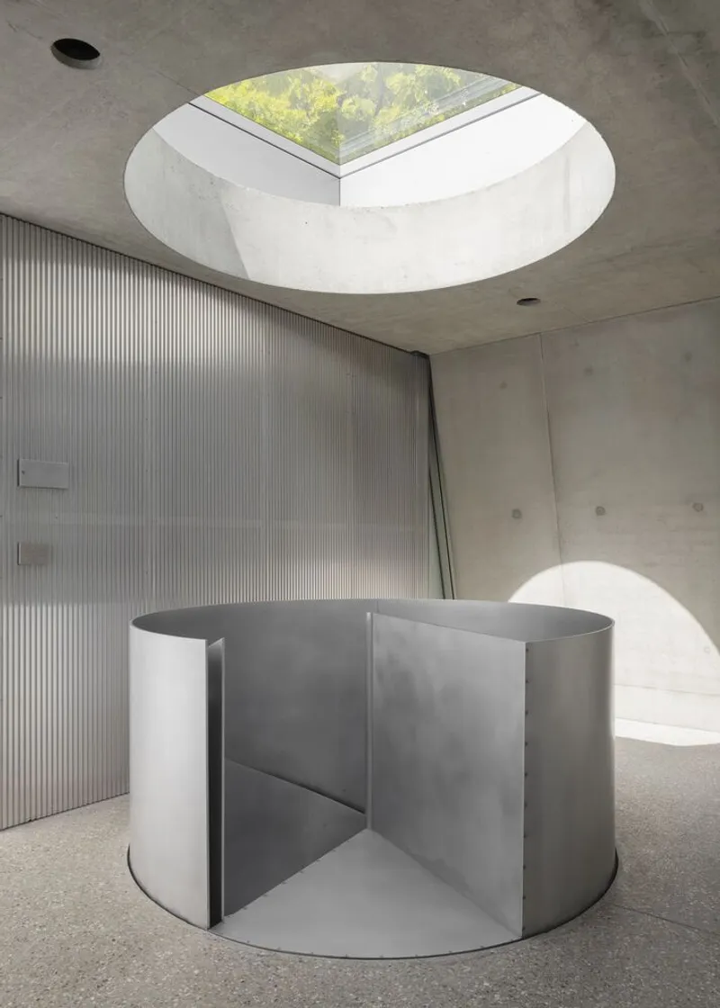 Stainless steel spiral stair with a circular oculus skylight above, concrete walls framing the vertical circulation.