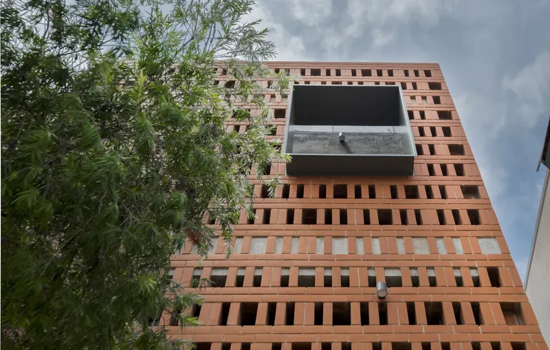 Looking up: brick jaali facade rising multiple storeys, single concrete cantilevered balcony with metal railing, bamboo at the edges