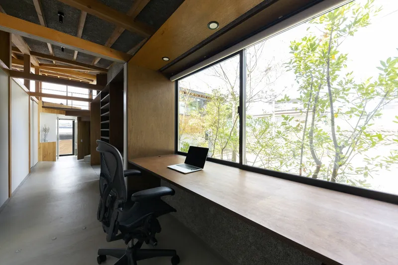 Study nook: long pale timber desk with two black ergonomic chairs, horizontal window framing a maple tree, dark timber wall, exposed ceiling
