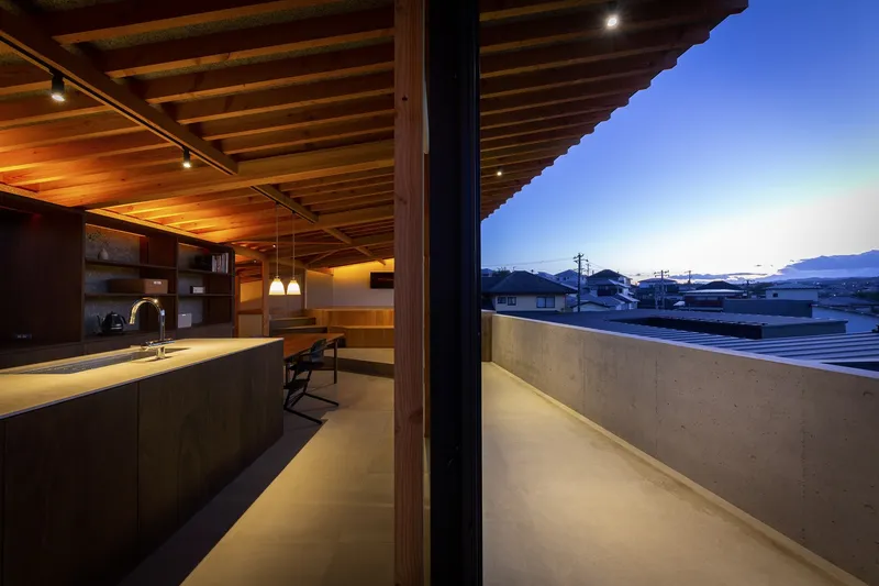 Interior at dusk: warm pendant lights over the kitchen counter and dining area, exposed rafter ceiling, sliding glass open to the balcony and distant hills