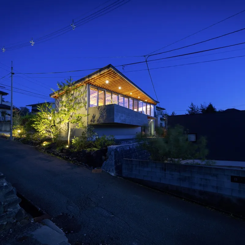 Street view at night: the house glowing warm on its slight rise above the road, timber rafter ceiling visible through the full-height glazing, power lines and trees in the foreground