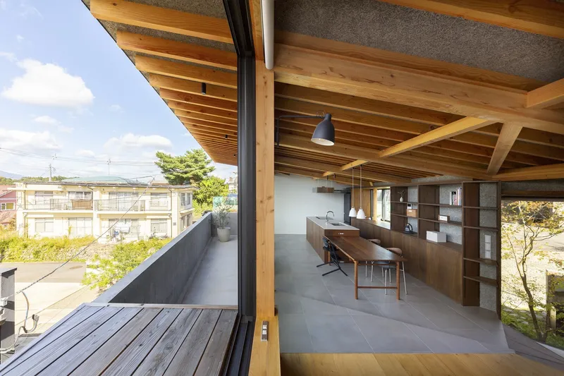 Upper living room: exposed timber rafter ceiling, long dining table, concrete floor, dark timber storage wall, full-height glass opening to the balcony with distant hills