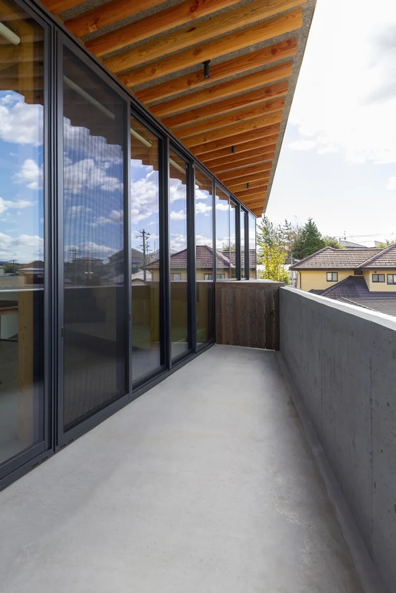 Balcony walkway: concrete floor and parapet, full-height glass sliders on the left, view across the neighbouring rooftops to distant hills