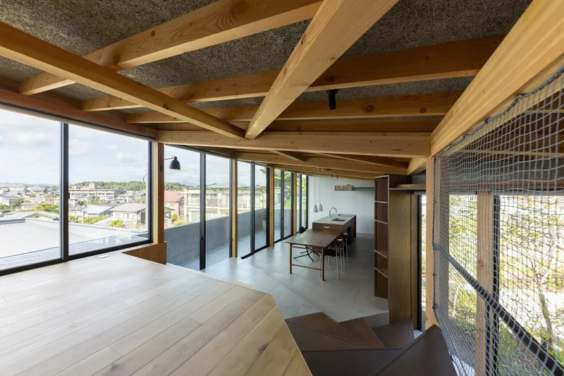 Upper open plan: dense timber rafter ceiling, dining table, concrete floor, wire mesh stair balustrade at right, full-width glazing framing the town view