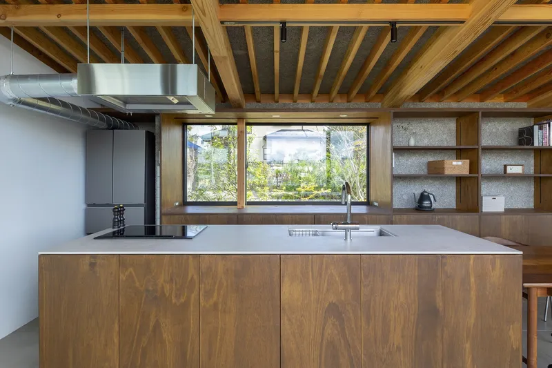 Kitchen island: dark timber base with pale stone counter, stainless steel extractor hood, horizontal window framing the garden, rafter ceiling, open timber shelving