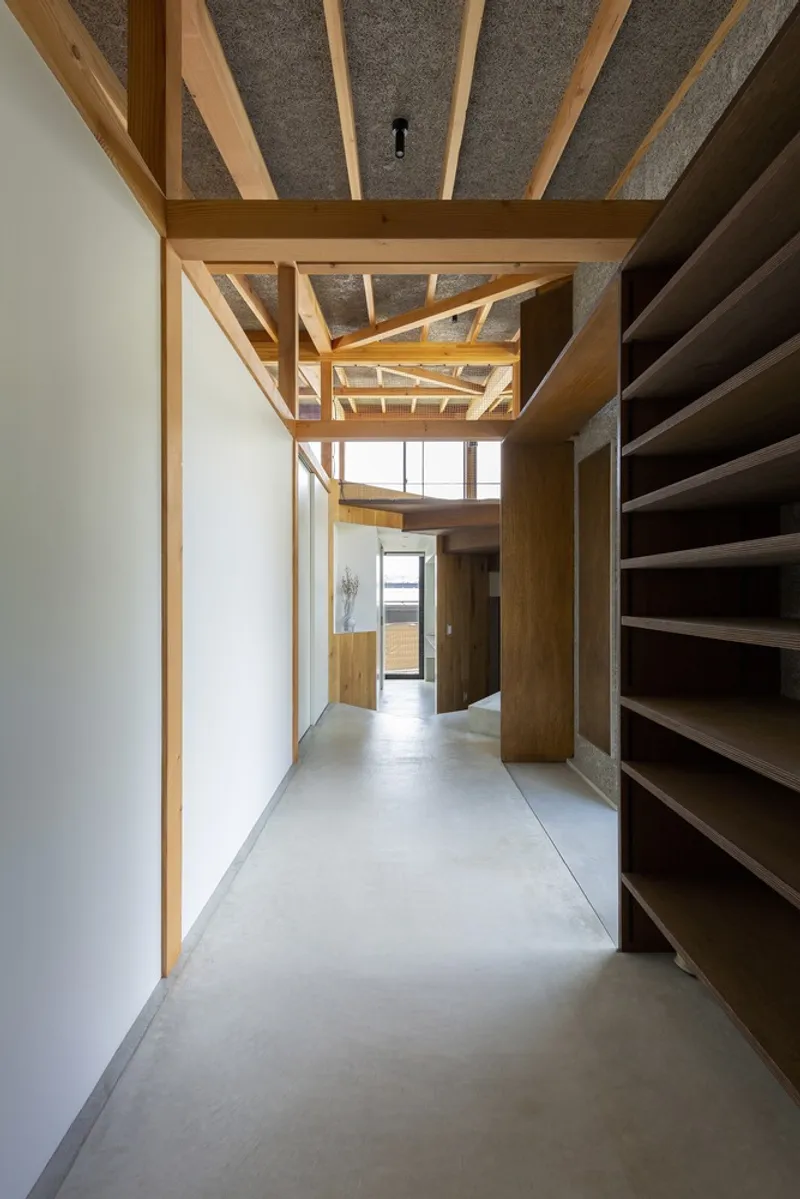 Corridor: timber shelving on the right, exposed rafter ceiling above, concrete floor, view through to a brighter room at the end