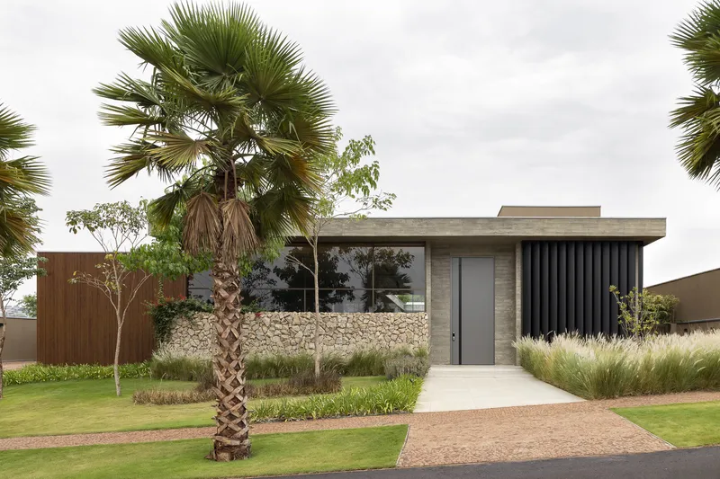 Street view of the single-story residence with vertical black slat screen and gabion wall behind palm trees
