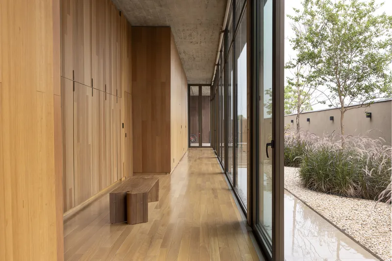 Timber-lined corridor with exposed concrete ceiling alongside planted courtyard with grasses beyond full-height glass