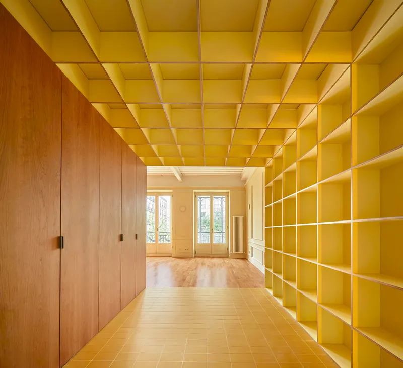 Yellow shelving wall and coffered ceiling in corridor leading to rooms with tall windows