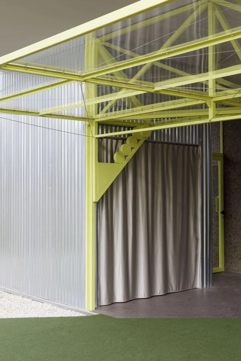 Detail of lime green steel truss supporting translucent wall panels beside silver curtain and artificial turf