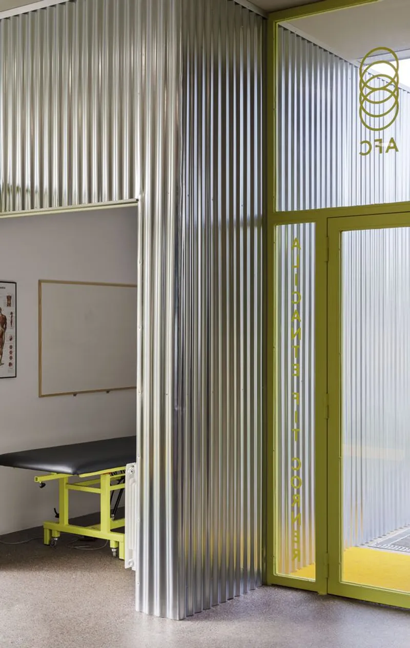 Interior partition of vertical corrugated metal panels with yellow steel frame and examination table visible