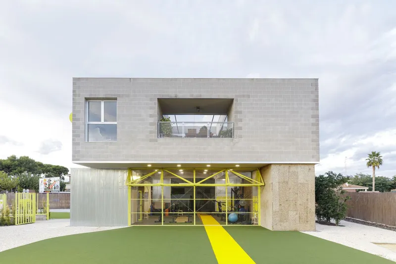 Street facade showing concrete block upper volume hovering above yellow steel-framed ground floor with corrugated metal cladding