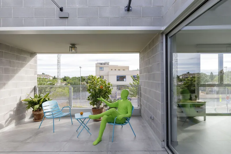 Covered terrace with concrete block walls and sliding glass doors, featuring a figure in green bodysuit seated among potted plants