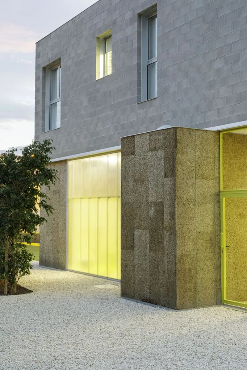 Ground floor entry illuminated at dusk with yellow-lit translucent panels behind cork-clad columns and concrete block upper level