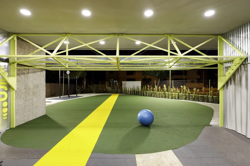 Covered play area with yellow steel truss structure over curved green turf path and blue exercise ball at night