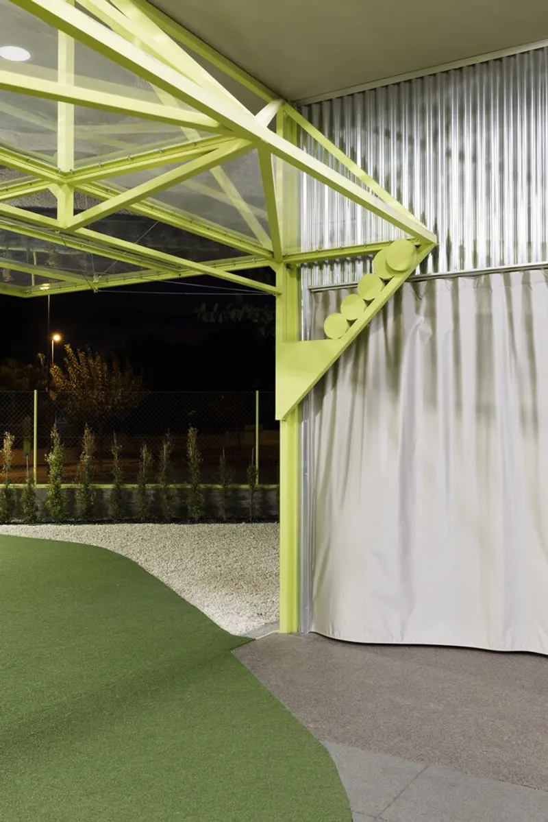 Covered entrance with exposed yellow truss framework and corrugated metal cladding at night