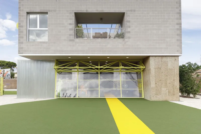 Daytime view of the concrete block upper volume cantilevering over yellow truss and green synthetic lawn