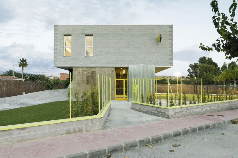 Street view of the two-storey block facade with yellow corrugated metal entry fence and planted beds