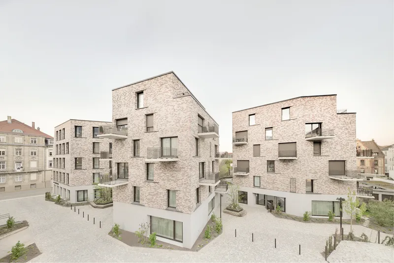 Street-level view of two light brick residential volumes with recessed balconies and planted courtyard below