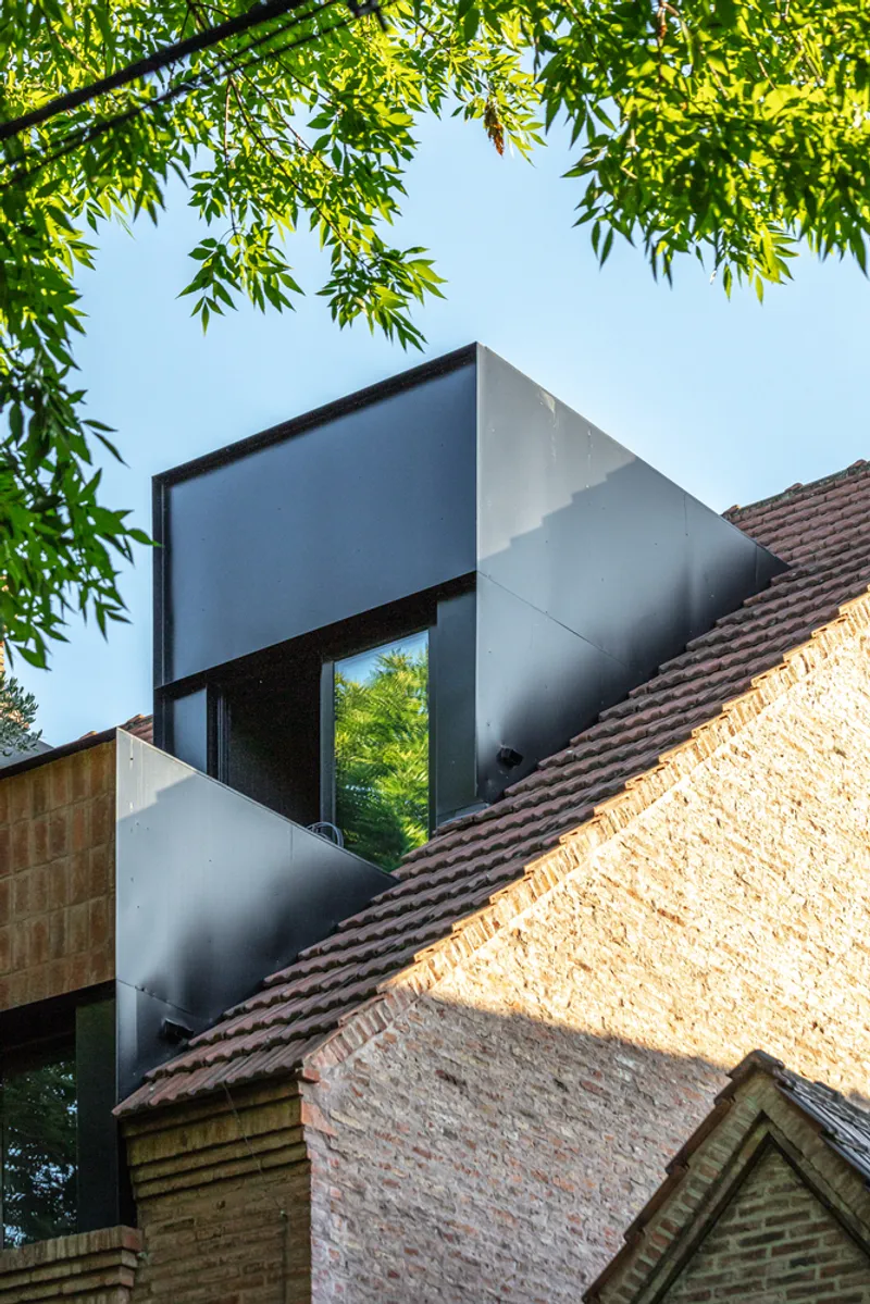 Metal-clad dormer addition rising above existing terracotta tile roofs framed by green foliage in daylight