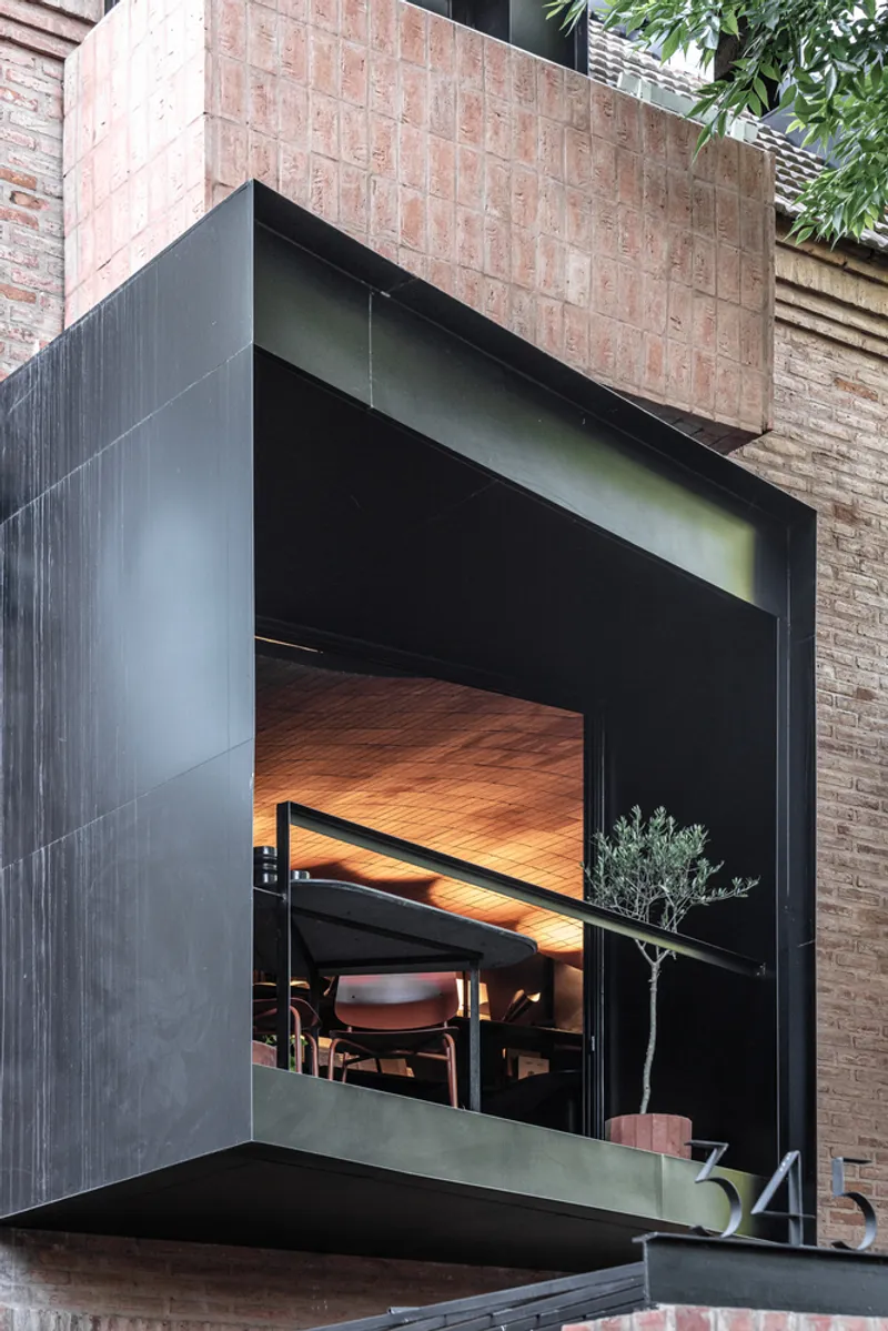 Cantilevered volume clad in dark metal panels with large glazed opening revealing wooden ceiling and dining furniture