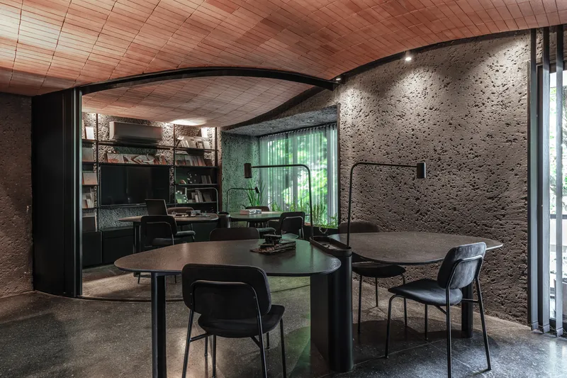 Interior dining space with textured concrete walls, timber plank ceiling and curved metal partition enclosing kitchen