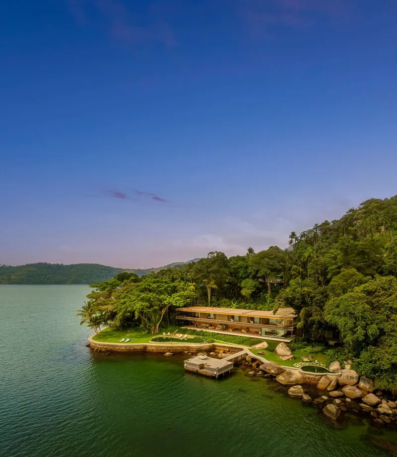 Aerial view of the horizontal timber-clad volume perched on a forested lakeside peninsula at dusk