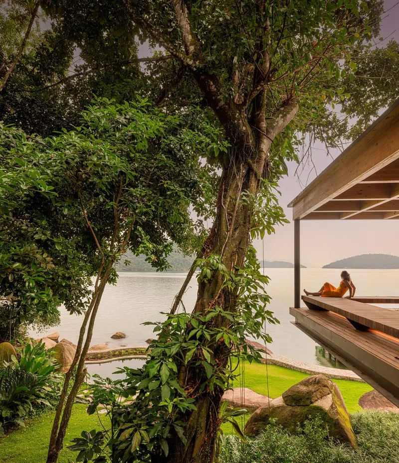 Cantilevered timber deck alongside a moss-covered boulder and reflecting pool beneath mature trees facing the water