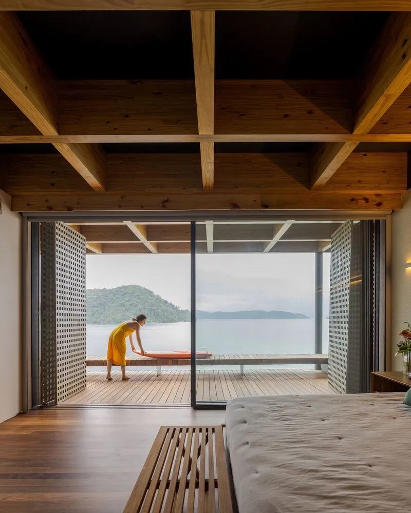 Timber-ceilinged bedroom opening onto a wooden deck with perforated metal screens framing lake and mountain views