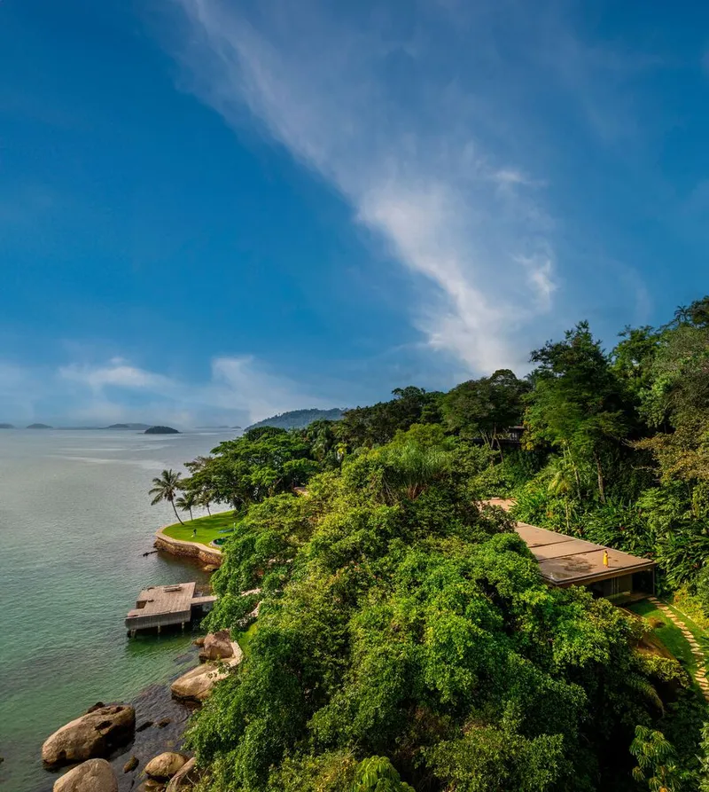 Aerial view of low-slung roof nestled into forested hillside above coastal waterfront