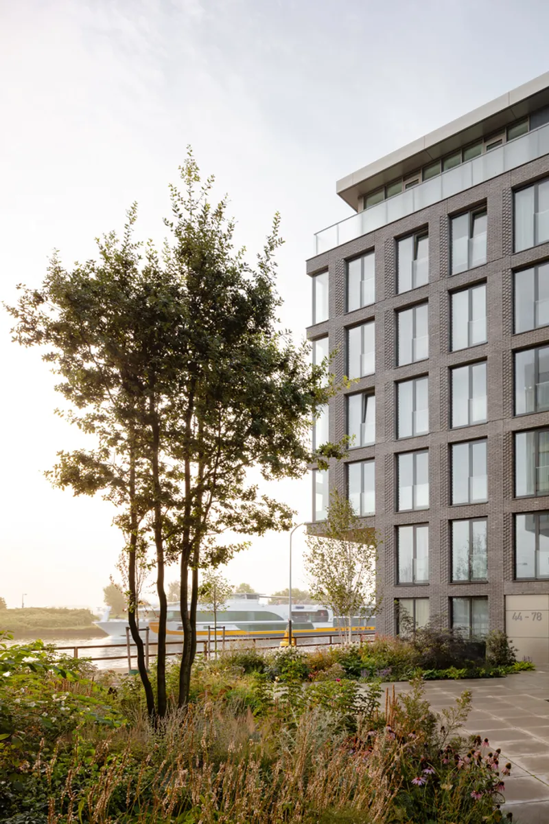 Corner view of the brick facade with large vertical windows beside a tree-lined waterfront path