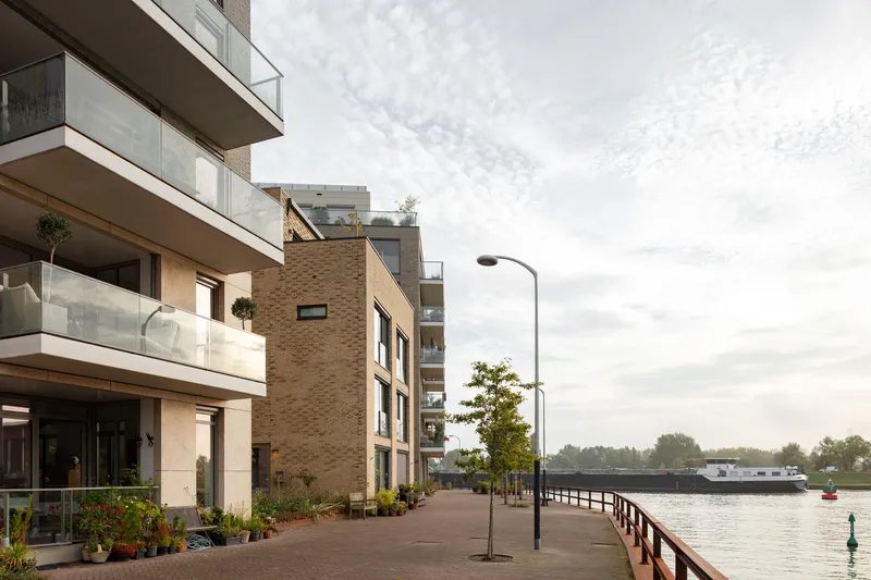 Riverside promenade with brick-clad residential blocks featuring stacked balconies and glass railings under cloudy skies