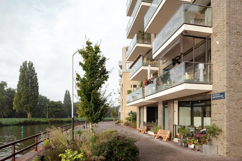 Waterside walkway with planted beds along brick residential facade with cantilevered balconies and glass railings