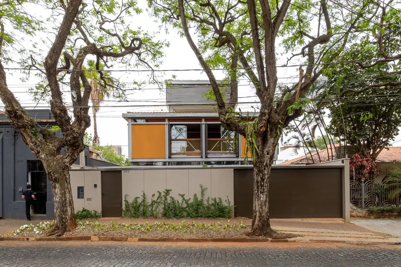 Street view of the raised upper volume with orange panels and large windows behind mature trees