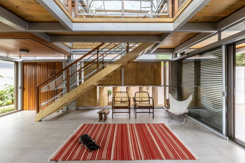 Double-height interior with timber and steel staircase ascending past a central skylight above a striped rug