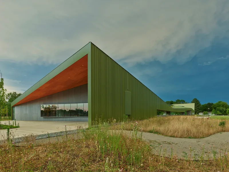 Green corrugated metal volume with a cantilevered wood soffit and ribbon glazing beside a native grass meadow