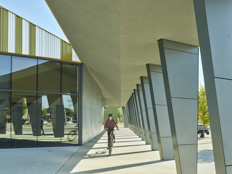 Covered walkway with angled metal columns and glass curtain wall as a cyclist passes beneath