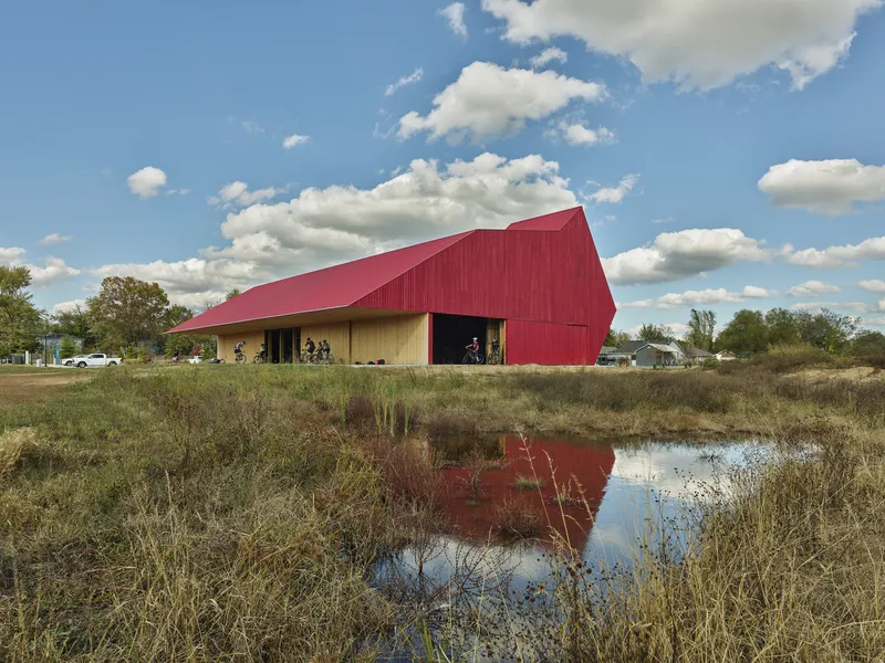 Red corrugated metal facade with angular roofline reflected in a pond beneath cumulus clouds