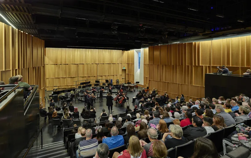 Concert hall with orchestra performing to seated audience beneath black ceiling and backlit timber acoustic panels