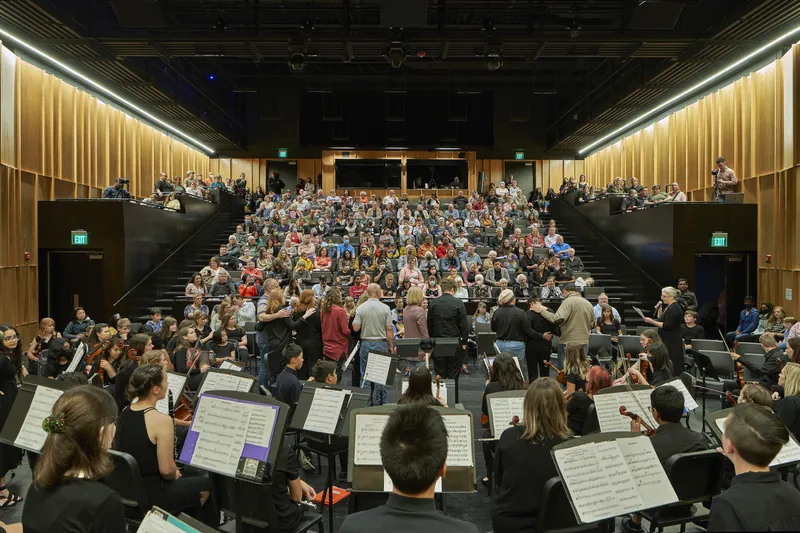 View from orchestra pit toward packed auditorium seating rising in tiered rows beneath timber acoustic wall panels