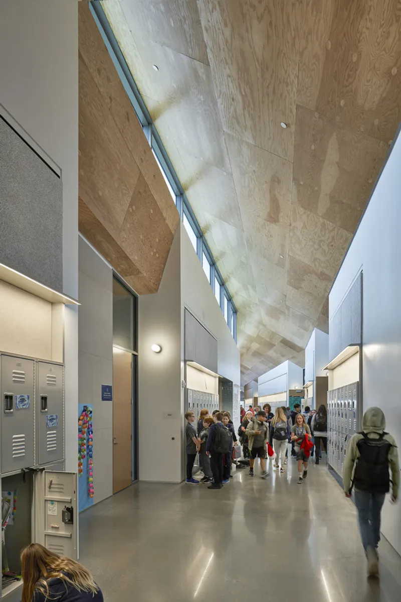 Interior corridor with exposed plywood ceiling, high clerestory windows, and students gathered along polished concrete floors