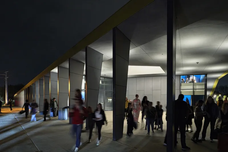 Covered exterior walkway with black metal columns and students moving past illuminated wall panels at night