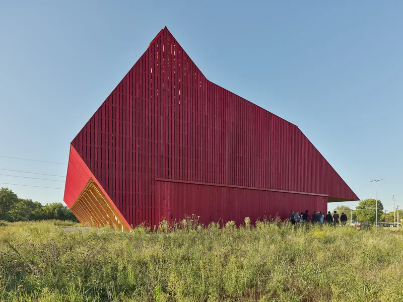 Gabled red metal volume with vertical cladding rising above tall grasses under clear sky
