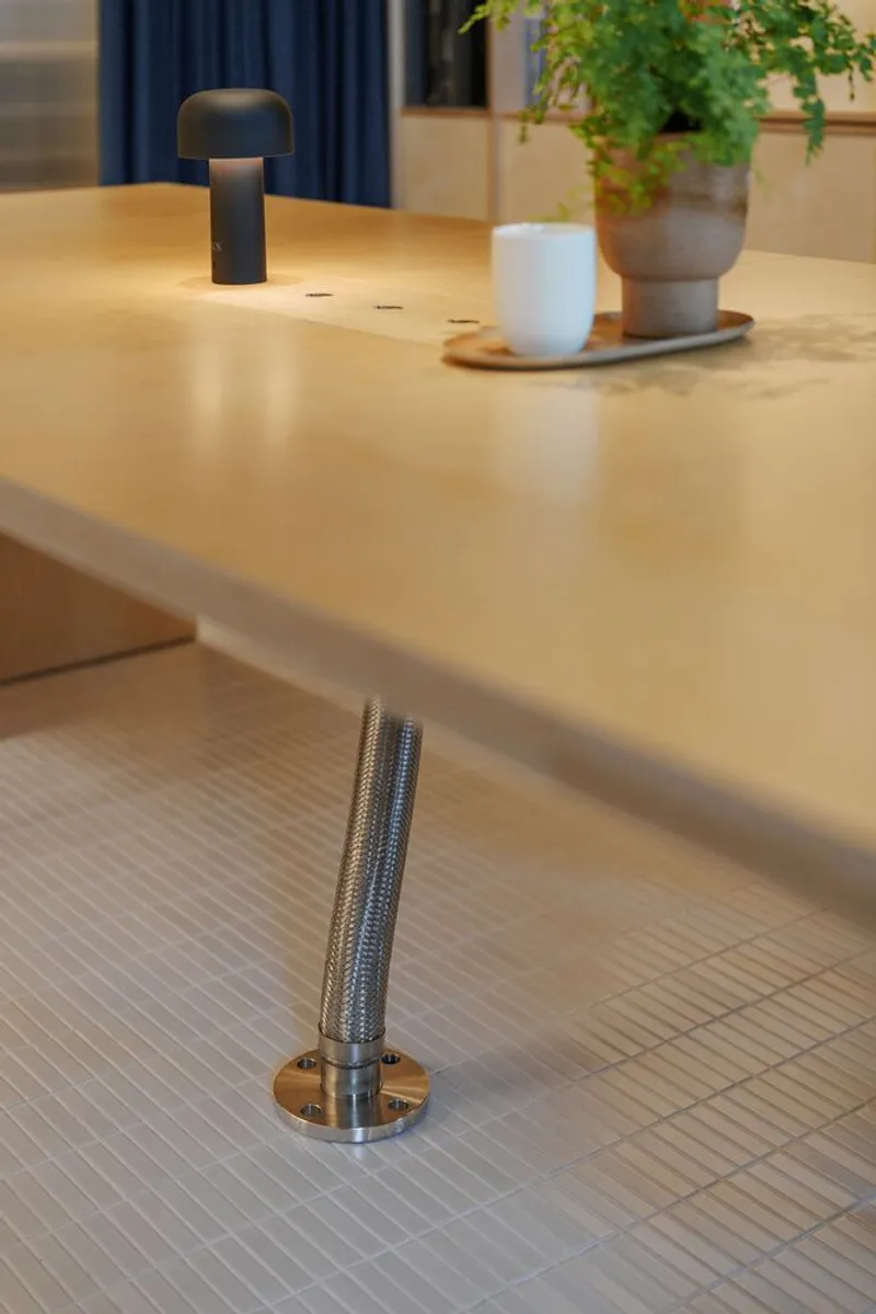 Plywood tabletop with knurled metal footrest rail anchored to white tiled floor beneath table edge