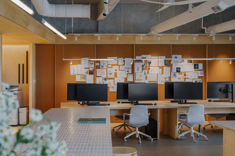 Office workspace with plywood workstations and pin-up wall under exposed ductwork and acoustic ceiling panels