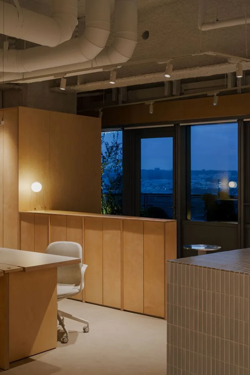 Office workspace with timber cabinetry and exposed concrete ceiling at dusk with city lights visible through glazing