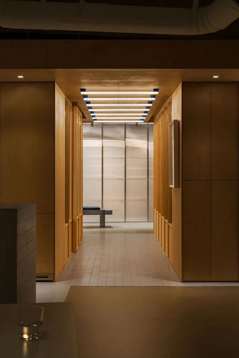 Corridor with parallel timber and fluted glass partitions leading to translucent screen lit by overhead linear fixtures