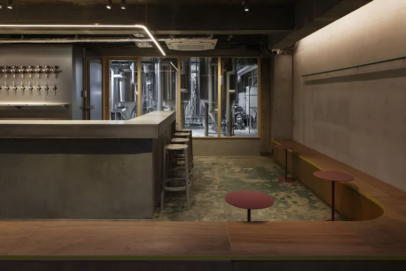Interior taproom with concrete bar, exposed brewery tanks behind glass, and round upholstered stools